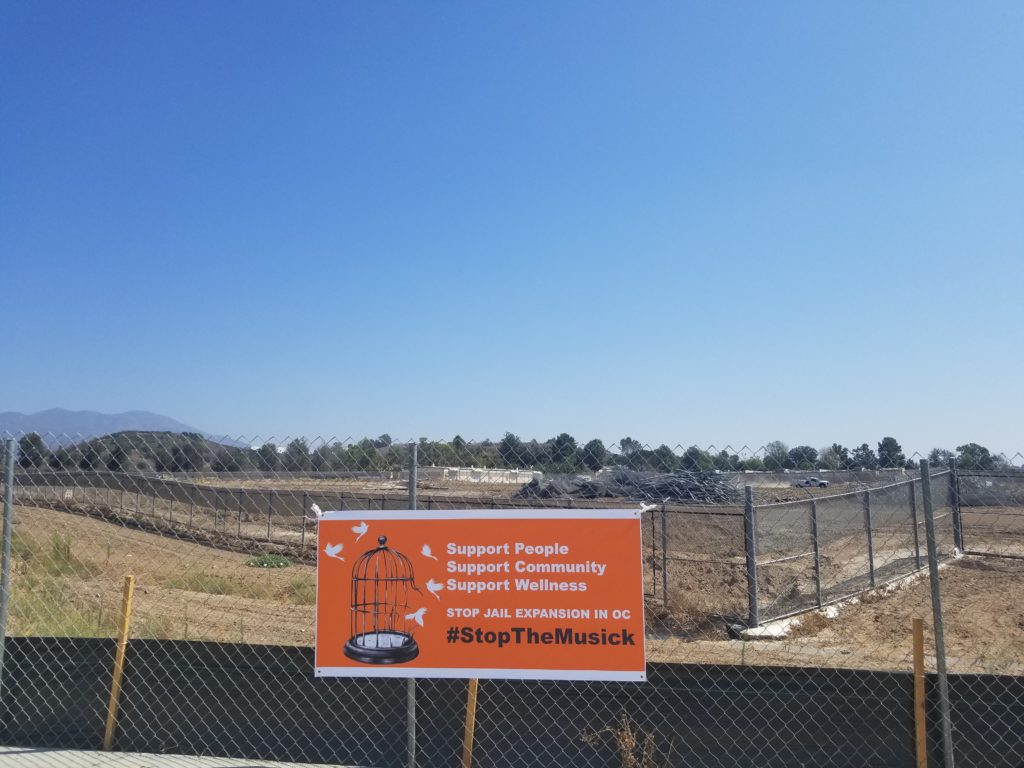 Photo of an orange banner hung on barbed wire fence in front of the Musick construction site. The banner has an image of white birds flying out of a broken bird cage and reads "Support People; Support Community; Support Wellness; Stop Jail Expansion in OC; #StopTheMusick."